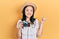 Young chinese girl wearing summer hat holding vintage camera smiling happy pointing with hand and finger to the side Royalty Free Stock Photo