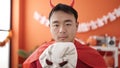 Young chinese man wearing devil costume holding skull mask at home Royalty Free Stock Photo