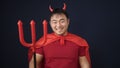Young chinese man wearing devil costume holding trident over isolated black background