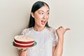 Young chinese woman holding carrot cake pointing thumb up to the side smiling happy with open mouth Royalty Free Stock Photo