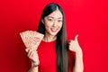 Young chinese woman holding mexican pesos smiling happy and positive, thumb up doing excellent and approval sign