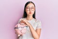 Young chinese woman holding piggy bank with glasses puffing cheeks with funny face