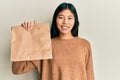 Young chinese woman holding take away paper bag looking positive and happy standing and smiling with a confident smile showing Royalty Free Stock Photo