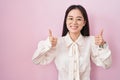 Young chinese woman standing over pink background success sign doing positive gesture with hand, thumbs up smiling and happy Royalty Free Stock Photo