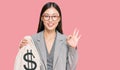 Young chinese woman wearing business suit holding dollars bag doing ok sign with fingers, smiling friendly gesturing excellent