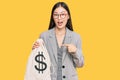 Young chinese woman wearing business suit holding dollars bag smiling happy pointing with hand and finger