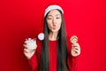 Young chinese woman wearing christmas hat holding cookies and milk making fish face with mouth and squinting eyes, crazy and Royalty Free Stock Photo