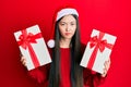 Young chinese woman wearing christmas hat and holding gifts skeptic and nervous, frowning upset because of problem