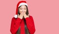 Young chinese woman wearing christmas hat laughing and embarrassed giggle covering mouth with hands, gossip and scandal concept