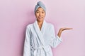 Young chinese woman wearing shower towel cap and bathrobe smiling cheerful presenting and pointing with palm of hand looking at