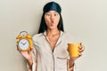 Young chinese woman wearing sleep mask and robe holding alarm clock and cup of coffee making fish face with mouth and squinting Royalty Free Stock Photo