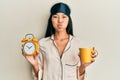 Young chinese woman wearing sleep mask and robe holding alarm clock and cup of coffee puffing cheeks with funny face Royalty Free Stock Photo