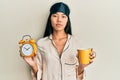 Young chinese woman wearing sleep mask and robe holding alarm clock and cup of coffee relaxed with serious expression on face Royalty Free Stock Photo