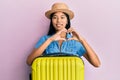 Young chinese woman wearing summer hat and holding cabin bag smiling in love doing heart symbol shape with hands