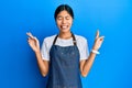 Young chinese woman wearing waiter apron gesturing finger crossed smiling with hope and eyes closed