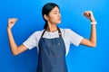 Young chinese woman wearing waiter apron showing arms muscles smiling proud