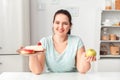 Making Right Choice. Chubby girl sitting at kitchen choosing between plate with desserts and apple looking camera Royalty Free Stock Photo