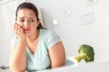 Body Care. Chubby girl sitting at kitchen table looking at broccoli unhappy close-up