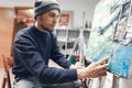 Young concentrated artist, sitting in a studio on a chair near the easel with the host, applying an oil paint using a knife