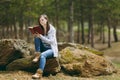 Young concentrated beautiful woman in casual clothes sitting on stone studying reading book in city park or forest on Royalty Free Stock Photo