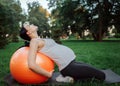 Young concentrated pregnant woman sit on knees on yoga mate in park. She lean to orange fitness ball with back and look