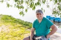 Young confident attractive man in casual dress and cap sitting and smiling at river or lake shore. Camping and travel Royalty Free Stock Photo