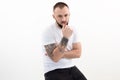 Young confident bearded man with short dark hair in white T-shirt, black jeans, sit, touching chin on white background. Royalty Free Stock Photo