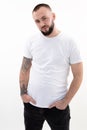 Young confident bearded man with short dark hair in white T-shirt, black jeans, standing, thrusting hands into pockets. Royalty Free Stock Photo