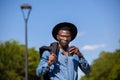 Young confident black man in a hat walking in a summer park holding a jacket over his shoulder and drinking coffee Royalty Free Stock Photo