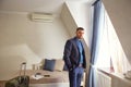 A young confident businessman in a formal suit stands in a hotel room. The suitcase is Packed for moving
