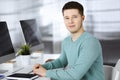 Young confident businessman, wearing a green pullover, is sitting at the desk in a modern office, while working on the Royalty Free Stock Photo