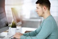 Young confident businessman, wearing a green pullover, is sitting at the desk in a sunny modern office, while working on Royalty Free Stock Photo