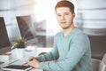 Young confident businessman, wearing a green pullover, is sitting at the desk in a sunny modern office, while working on Royalty Free Stock Photo