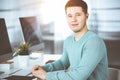 Young confident businessman, wearing a green pullover, is sitting at the desk in a sunny modern office, while working on Royalty Free Stock Photo