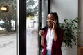 Young confident businesswoman standing in office looking through the window using smartphone or mobile phone for communication.