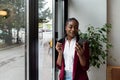 Young confident businesswoman standing in office looking through the window using smartphone or mobile phone for communication.