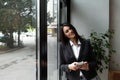 Young confident businesswoman standing in office looking through the window using smartphone or mobile phone for communication.