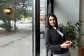 Young confident businesswoman standing in office looking through the window using smartphone or mobile phone for communication.