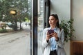 Young confident businesswoman standing in office looking through the window using smartphone or mobile phone for communication.