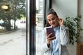 Young confident businesswoman standing in office looking through the window using smartphone or mobile phone for communication.