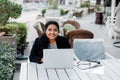 A young and confident Indian Asian woman smiles while sitting and working on her laptop in a stylish cafe on the street Royalty Free Stock Photo