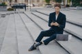 Young confident man in stylish suit using smartphone while sitting on steps outside with laptop Royalty Free Stock Photo