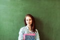 Young confident smiling female high school student standing in front of chalkboard in classroom, holding textbooks.