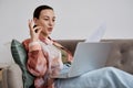 Young confident tutor with paper sitting on couch with laptop on her knees