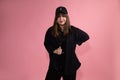 Young, confident woman dancer in black baseball cap, shirt and sportswear on pink background in studio looks confidently into