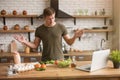 Young confused man standing in well-equipped kitchen has no idea what to cook for breakfast checking recipe in his laptop , sunday Royalty Free Stock Photo