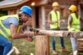 Young constructer worker, working with drilling machine on wooden construction. Other workers having a conversation in the