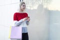 young consumer woman standing by a fashion store window display holding paper shopping bags and using a smartphone Royalty Free Stock Photo