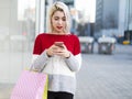 young consumer woman standing by a fashion store window display holding paper shopping bags and using a smartphone Royalty Free Stock Photo