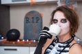 Young contented woman with creepy makeup and in skeleton costume drinks refreshing, relaxing or hot beverage from mug at Royalty Free Stock Photo